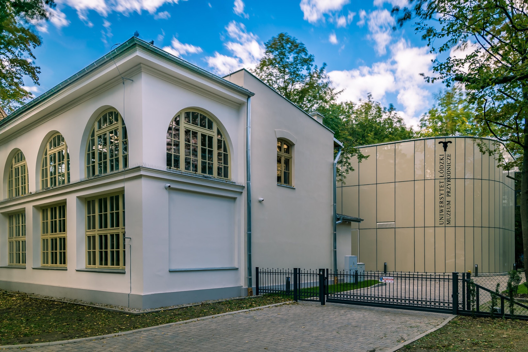 Odrestaurowany budynek Muzeum Przyrodniczego w Łodzi z charakterystycznymi łukowymi oknami i nowoczesną dobudówką, otoczony zielenią i ogrodzeniem.