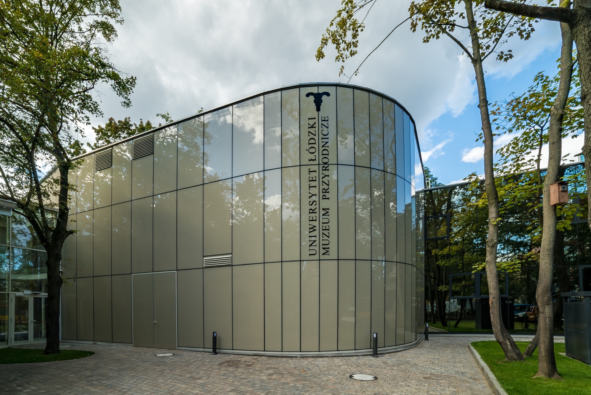 Nowoczesny budynek Muzeum Przyrodniczego Uniwersytetu Łódzkiego z przeszkloną fasadą, odbijającą drzewa i niebo, widziany z poziomu chodnika z kostki brukowej.