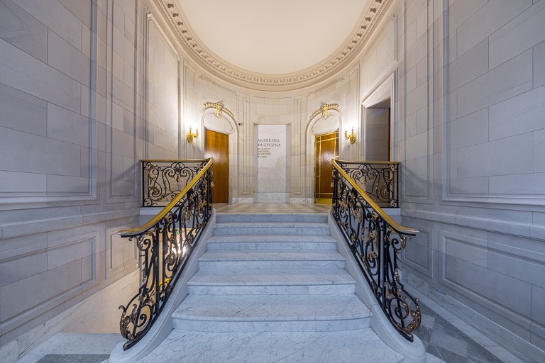 Marmurowe schody z ozdobnymi, złoconymi balustradami w budynku Akademii Muzycznej. Klasycystyczne wnętrze z kamiennymi ścianami i symetrycznym układem drzwi.