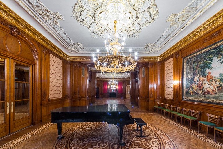 Wnętrze sali balowej z parkietem w ornamenty, drewnianymi boazeriami, żyrandolem i fortepianem. Widoczny fragment gobelinu i rzędu krzeseł.