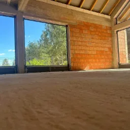 Wnętrze surowego domu w trakcie budowy, z zamontowanymi dużymi oknami z czarnymi ramami, widok na zewnątrz przez szyby, ściany z czerwonej cegły i betonowy strop.