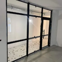 Szklana ściana działowa z czarnymi profilami i drzwiami, widoczne elementy wentylacji pod sufitem, podłoga wyłożona płytkami w dwóch wzorach.