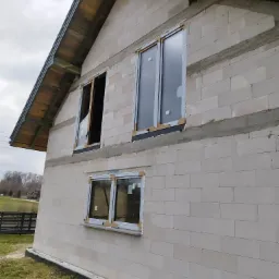 Budynek w trakcie montażu okien, widoczne trzy okna z zamontowanymi ramami, jedno bez szyby, surowy stan elewacji z betonu komórkowego.