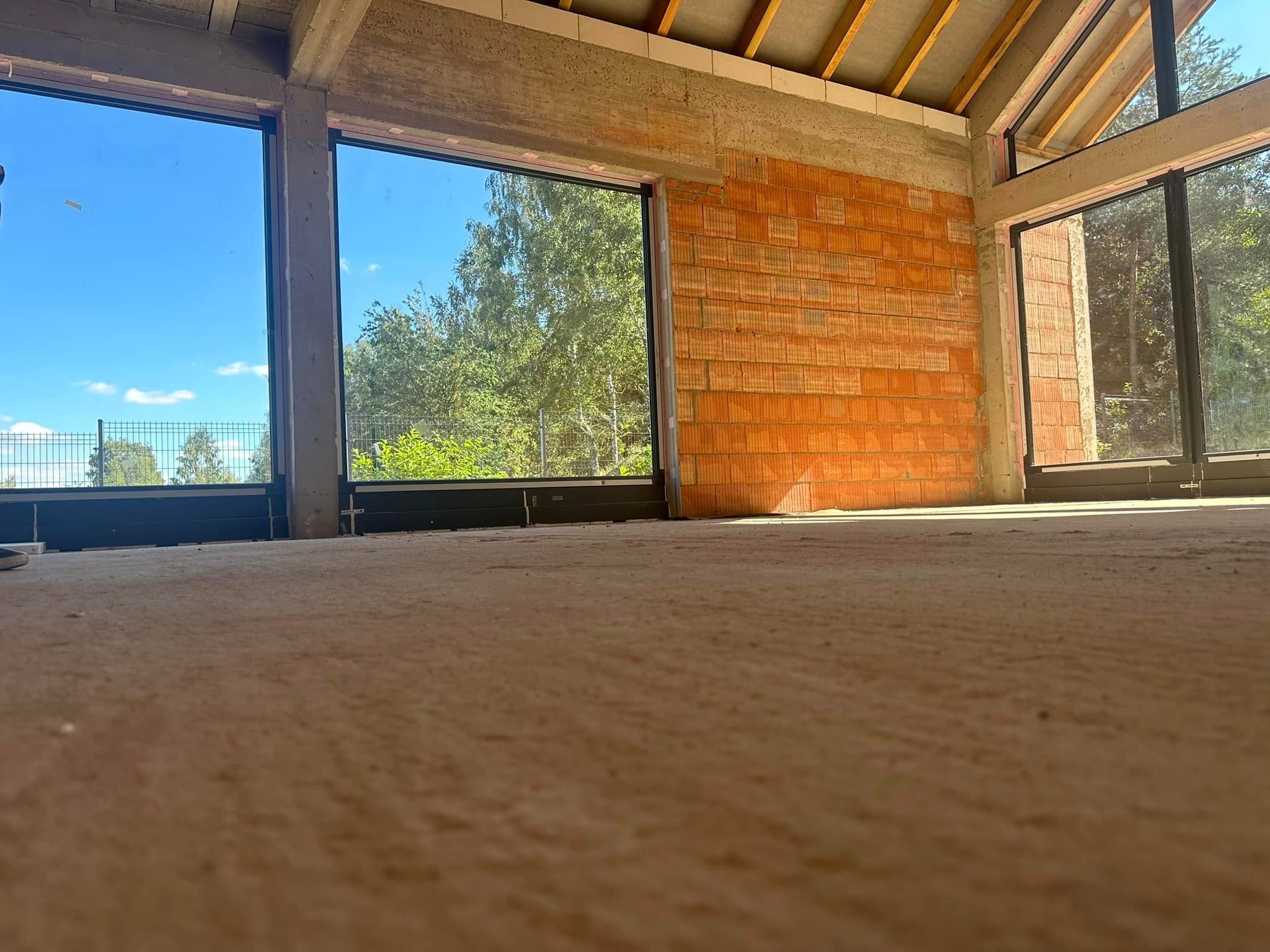 Wnętrze surowego domu w trakcie budowy, z zamontowanymi dużymi oknami z czarnymi ramami, widok na zewnątrz przez szyby, ściany z czerwonej cegły i betonowy strop.