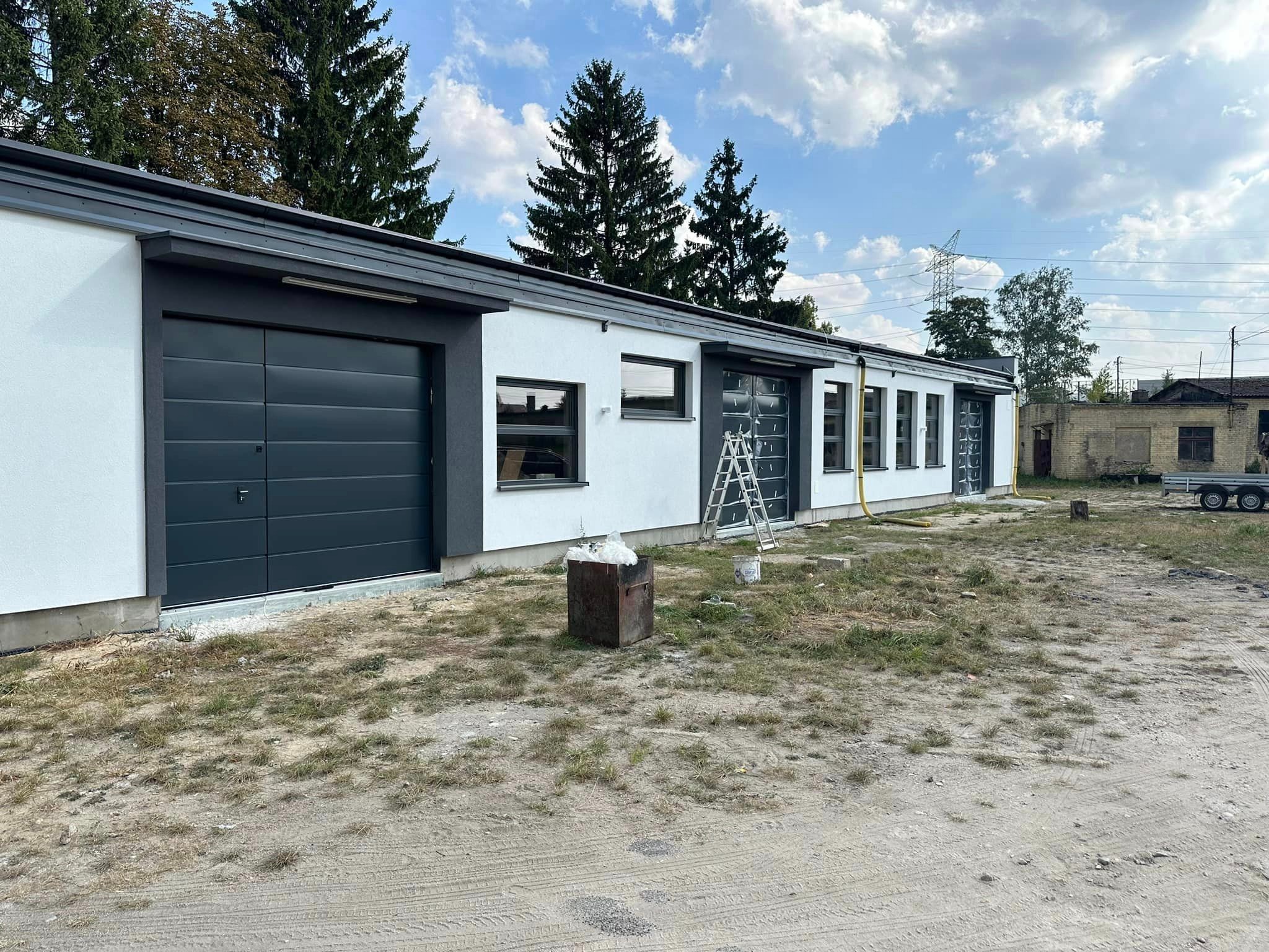 Nowoczesny budynek z szarymi bramami garażowymi segmentowymi, białymi ścianami i oknami, drabina oparta o ścianę, teren wokół budynku częściowo zagospodarowany.