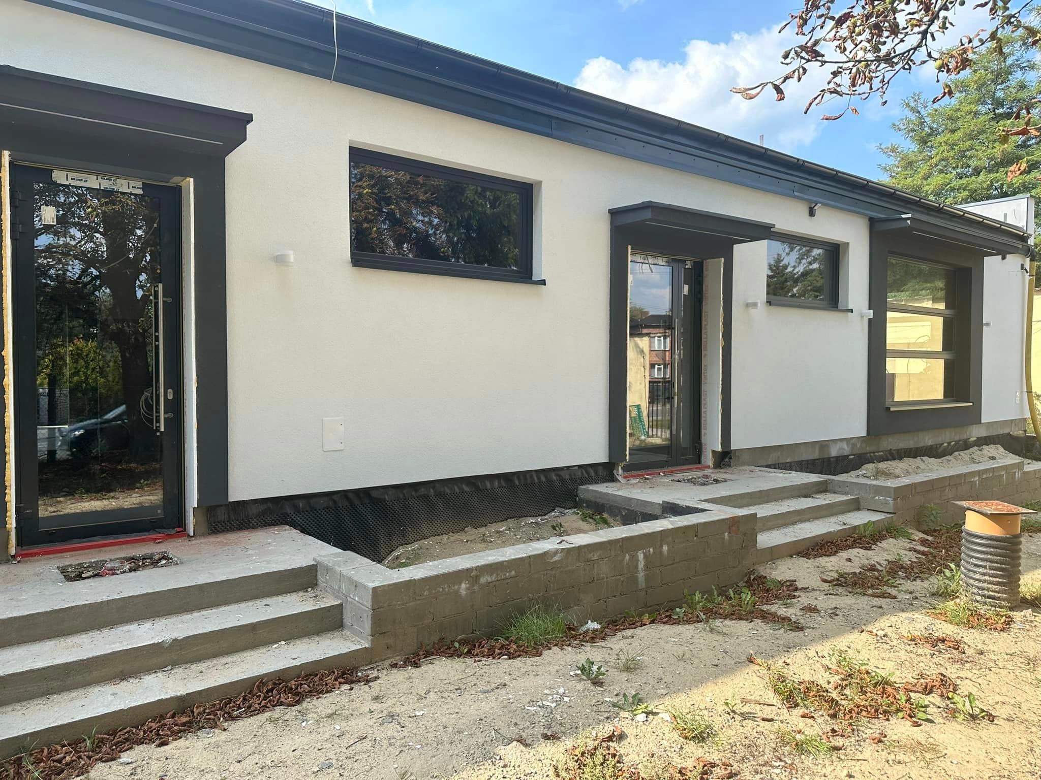 Nowoczesny dom z białymi ścianami i antracytowymi oknami oraz drzwiami zewnętrznymi, widoczne schody wejściowe w trakcie budowy, teren wokół domu niezagospodarowany, widoczne elementy izolacji...