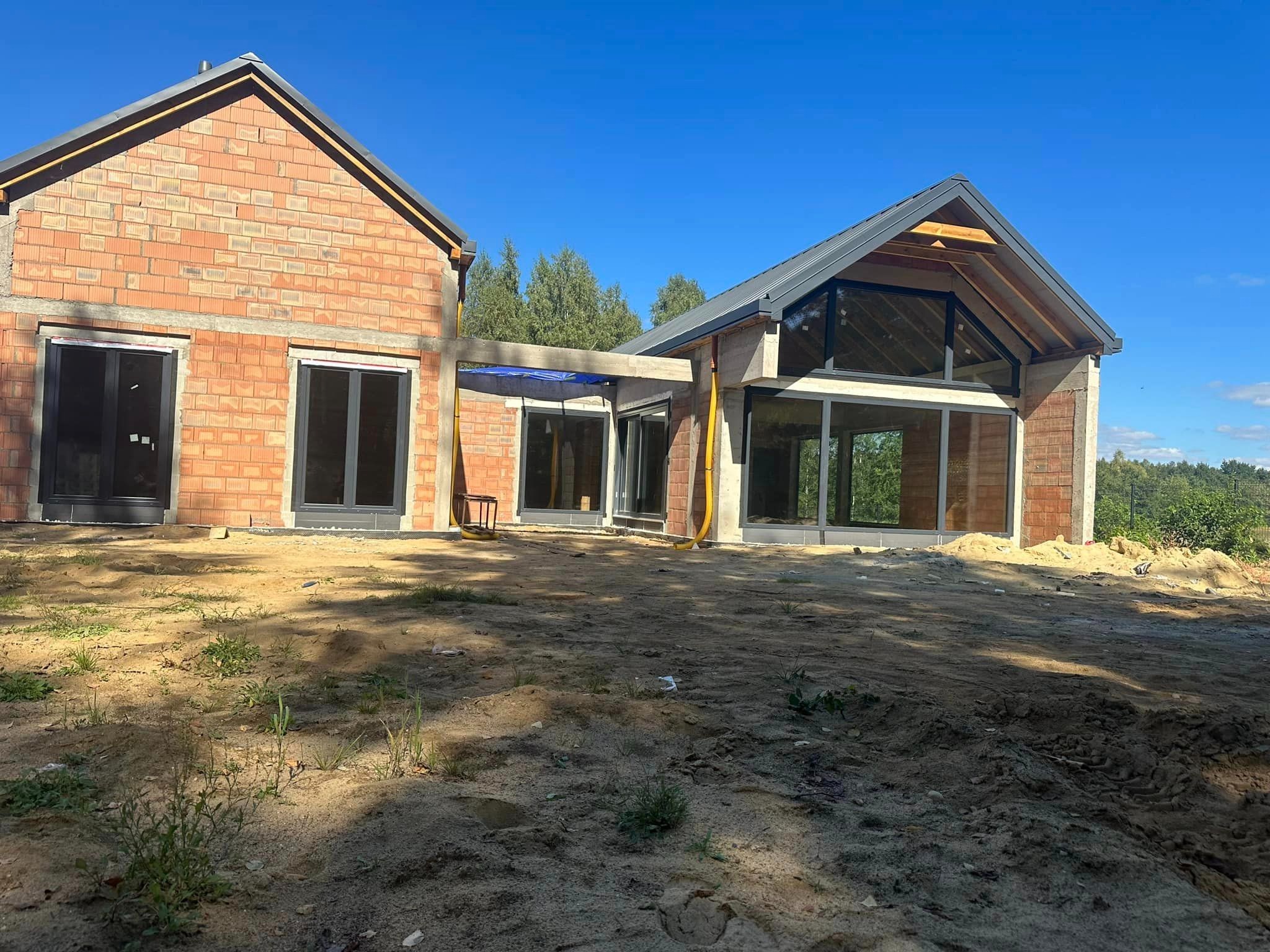 Surowy stan budowy domu jednorodzinnego z widocznymi oknami i niezadaszonym łącznikiem, teren wokół w trakcie niwelacji.