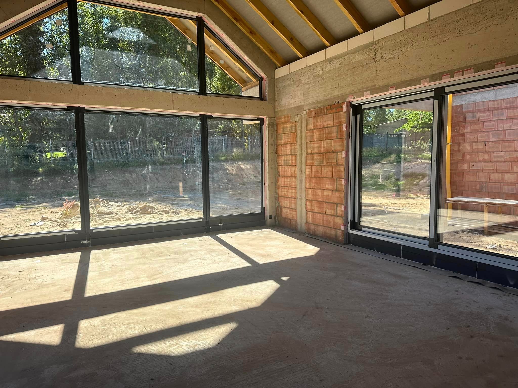 Wnętrze surowego budynku w Głownie z zamontowanymi oknami aluminiowymi o czarnych ramach, widoczne promienie słoneczne padające na betonową podłogę oraz częściowo wykończone ściany z cegły.