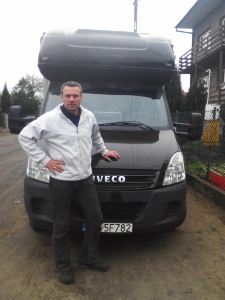 Mężczyzna opiera się o czarnego busa Iveco z bagażnikiem dachowym na tle domu jednorodzinnego. Widoczna tablica rejestracyjna 5F782.