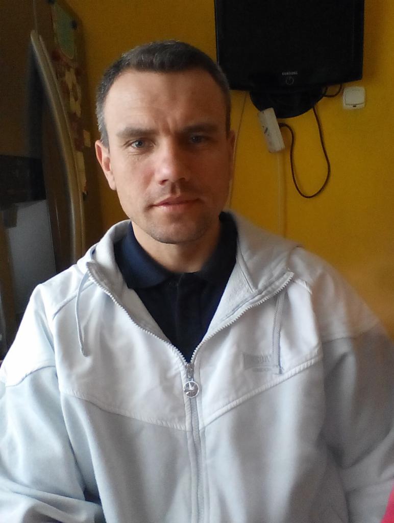 Portret mężczyzny w jasnej bluzie na tle żółtej ściany z telewizorem. Widoczne zmęczenie na twarzy, krótko ostrzyżone włosy i ciemna koszulka polo.