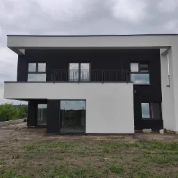 Dwukondygnacyjny dom z płaskim dachem, wykończony w czerni i bieli, z balkonem z metalową balustradą, na działce w trakcie zagospodarowania.