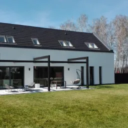 Nowoczesny, biały dom z ciemnym dachem i pergolą na tarasie. Widoczny zadbany trawnik i ogrodzenie. Jasne, naturalne światło słoneczne.