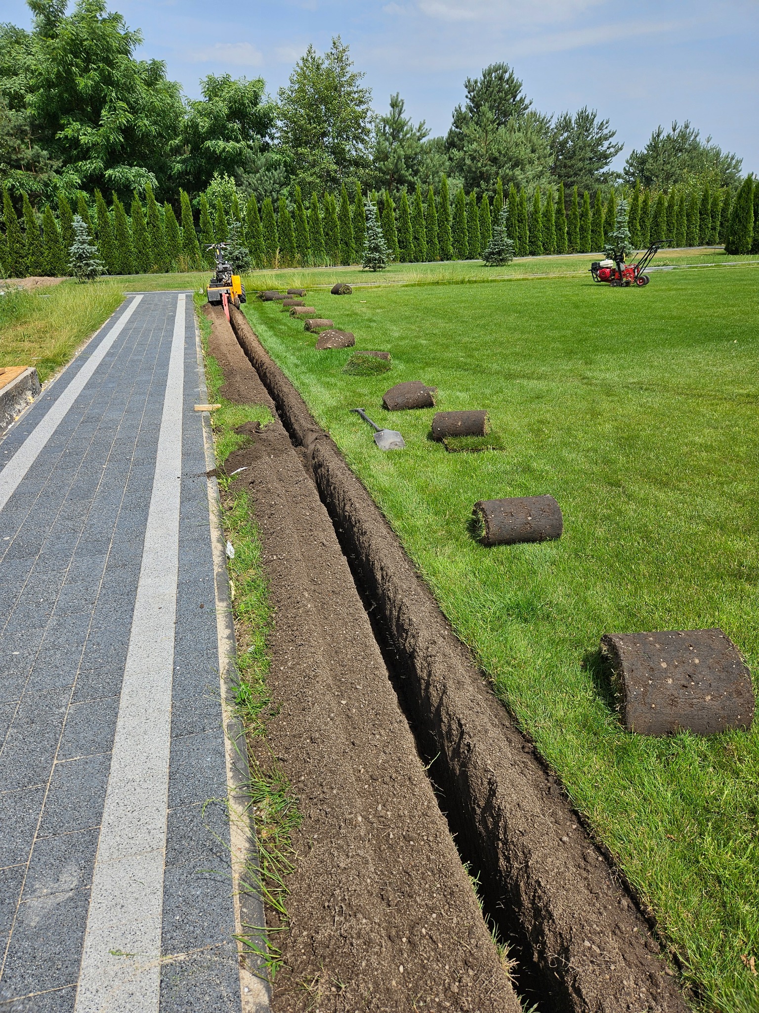 Przygotowanie terenu pod trawnik z rolki: wykopany rów, rozłożone rolki trawy, maszyna do cięcia darni w tle, obok ścieżka z kostki brukowej, zadbany trawnik z drzewami w tle.