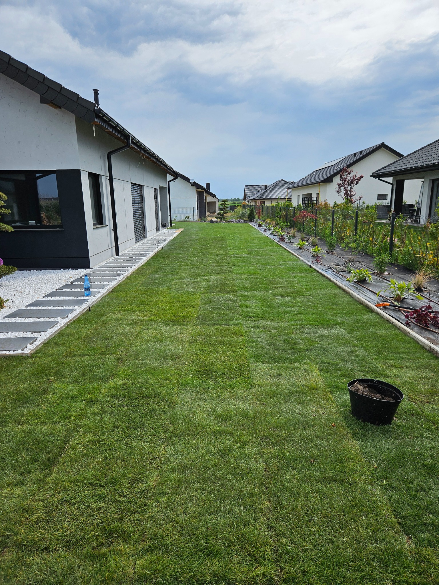 Długi, równo skoszony trawnik między nowoczesnymi domami, z ścieżką z płyt i białego kamienia po lewej stronie oraz rabatą z młodymi roślinami po prawej, widok perspektywiczny.