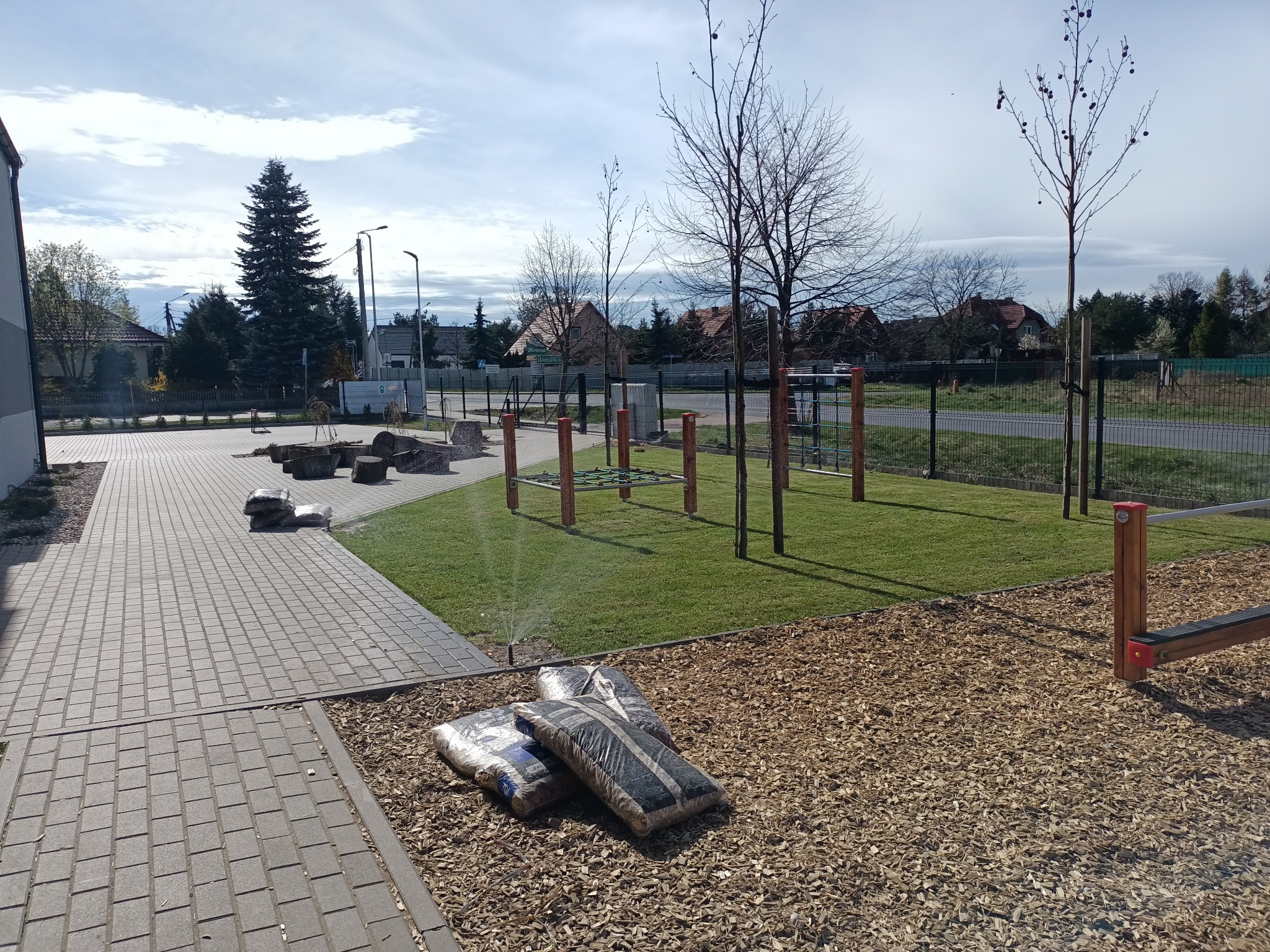 Nowo założony trawnik z systemem nawadniania, częściowo wyłożony korą, widoczne worki z podłożem, drzewa i elementy placu zabaw.