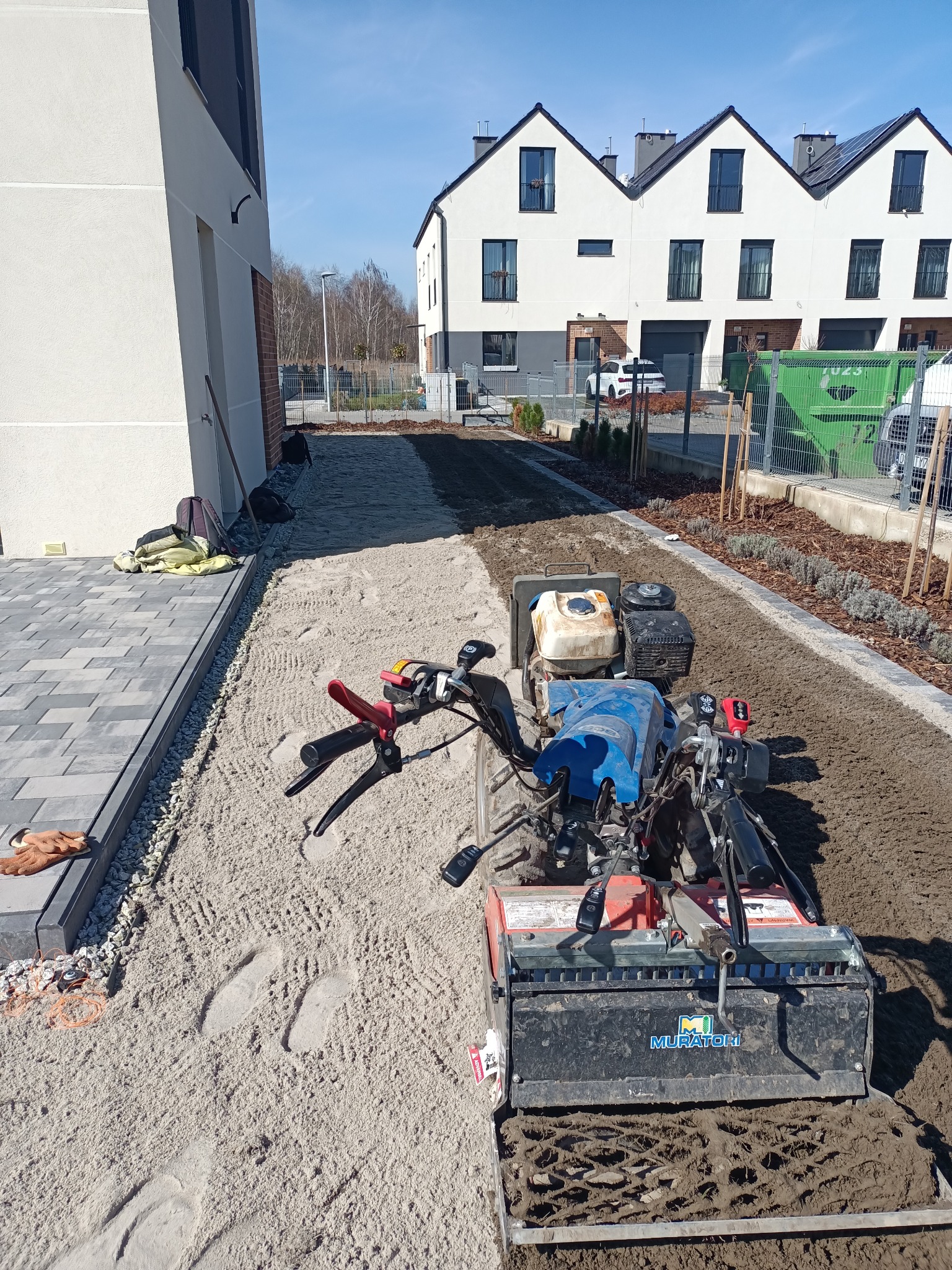 Glebogryzarka marki Muratori przygotowuje teren pod trawnik na piaszczystym podłożu, widoczne świeże ślady stóp i budynki w tle.