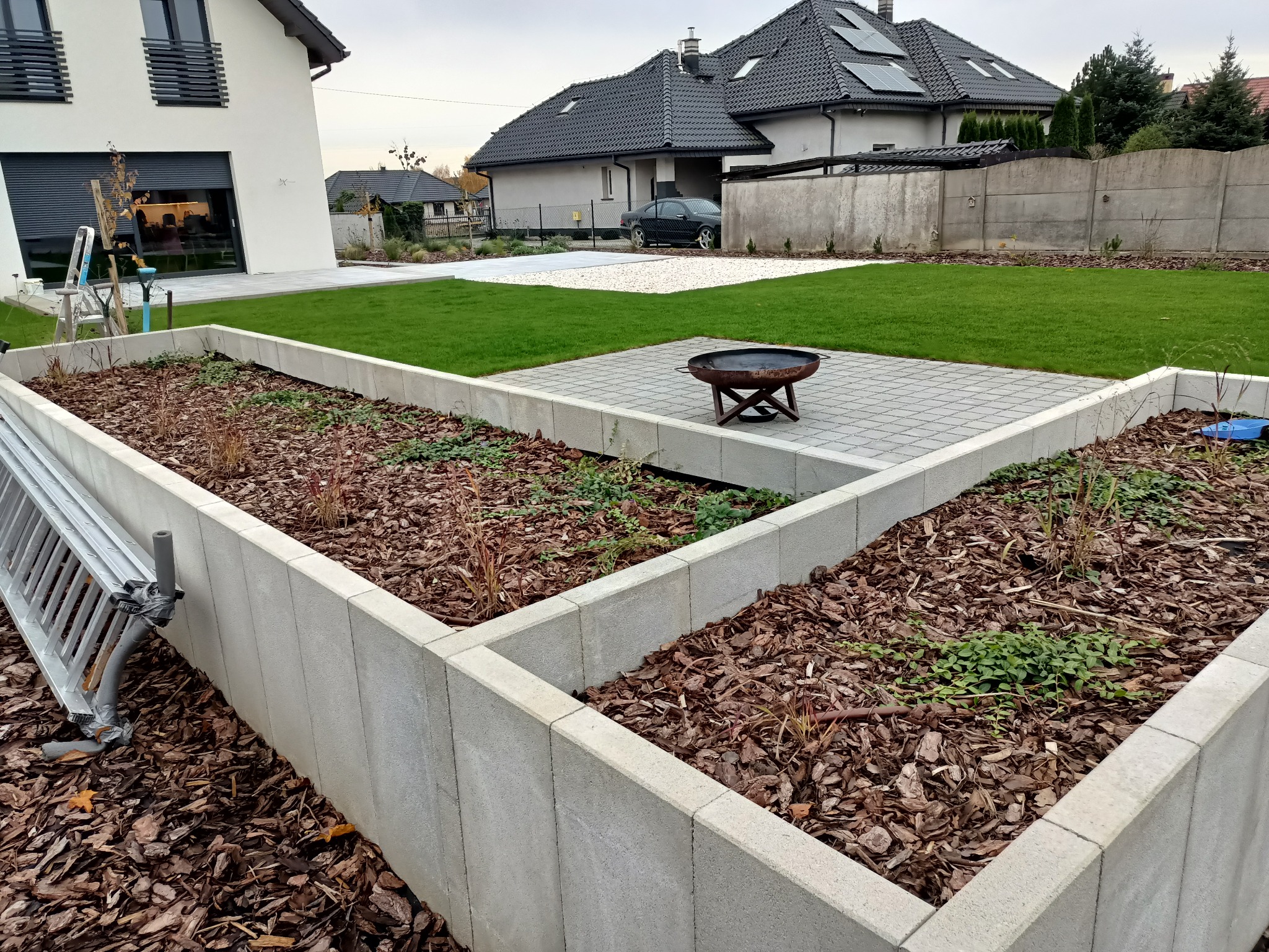 Nowoczesny ogród z podwyższonymi rabatami wyłożonymi korą, równo przystrzyżonym trawnikiem i paleniskiem na patio z kostki brukowej.