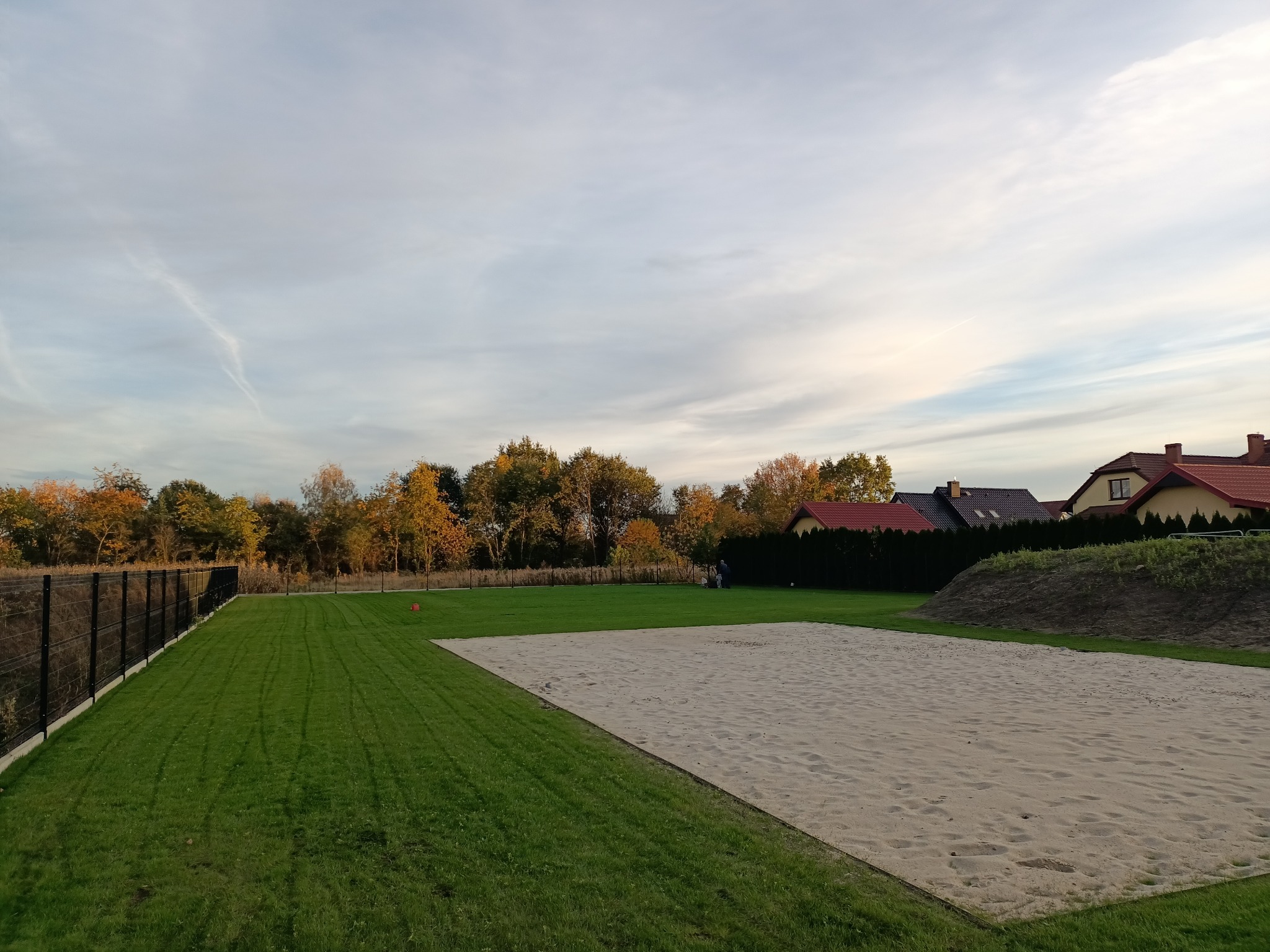 Przygotowany teren pod przyszły ogród z piaskowym podłożem, świeżo skoszonym trawnikiem i ogrodzeniem panelowym w tle, częściowo widoczne domy jednorodzinne.