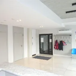 Jasne, minimalistyczne wnętrze biura z numerowanymi drzwiami, wieszakiem na ubrania i błękitnymi krzesłami. Marmurowe płytki podłogowe i nowoczesne oświetlenie.