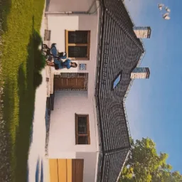 Wizualizacja domu jednorodzinnego z dwuspadowym dachem, widoczny fragment ogrodu, podjazd i osoba siedząca na krześle przed wejściem.