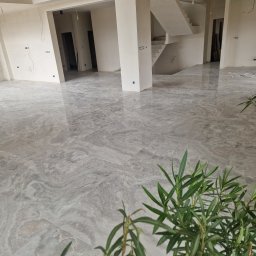 GUS ER-House Rafał Siwiec - Wnętrze w stanie deweloperskim z połyskującą, szarą podłogą imitującą marmur, widoczne schody, surowe ściany i roślina doniczkowa w pierwszym planie.