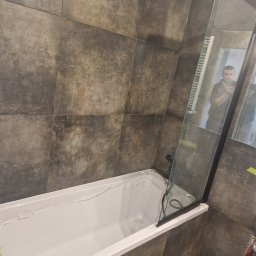 GUS ER-House Rafał Siwiec - Nowoczesna łazienka z wanną i szklanym parawanem, wykończona płytkami w odcieniach szarości. Widoczne detale wykończeniowe i odbicie osoby w lustrze.