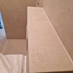 GUS ER-House Rafał Siwiec - Beżowa, kamienna zabudowa w łazience z widocznymi fugami i fragmentem niezapakowanego wyposażenia. Minimalistyczny design, ciepłe oświetlenie.