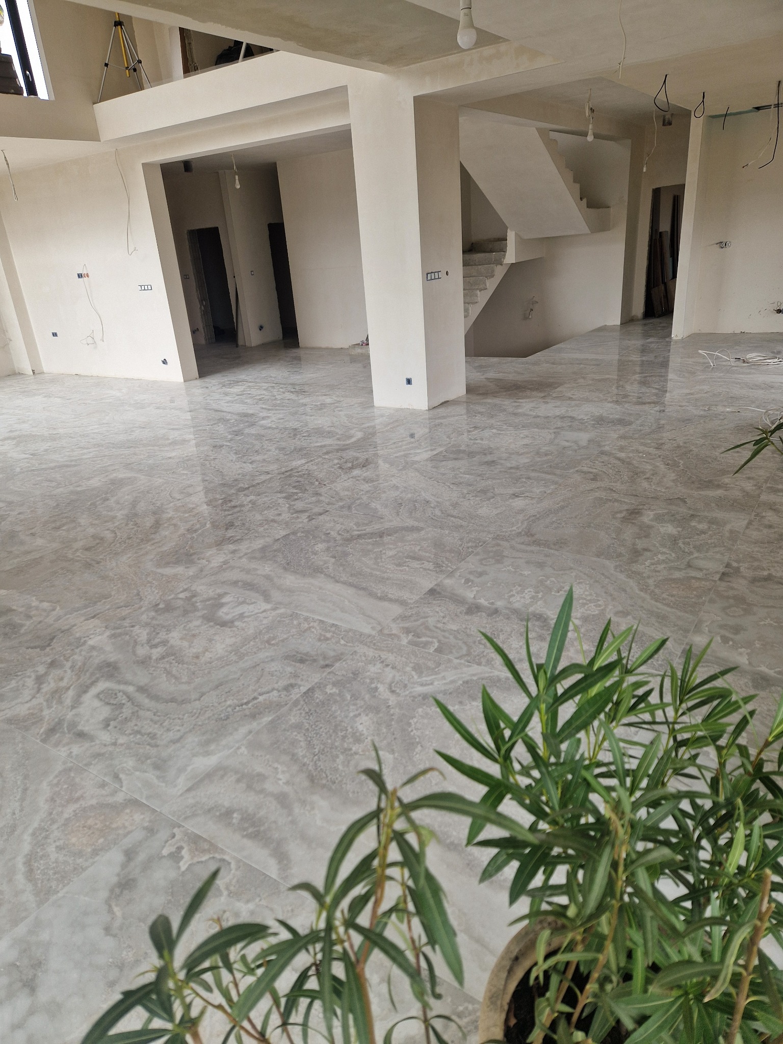 Wnętrze w stanie deweloperskim z połyskującą, szarą podłogą imitującą marmur, widoczne schody, surowe ściany i roślina doniczkowa w pierwszym planie.