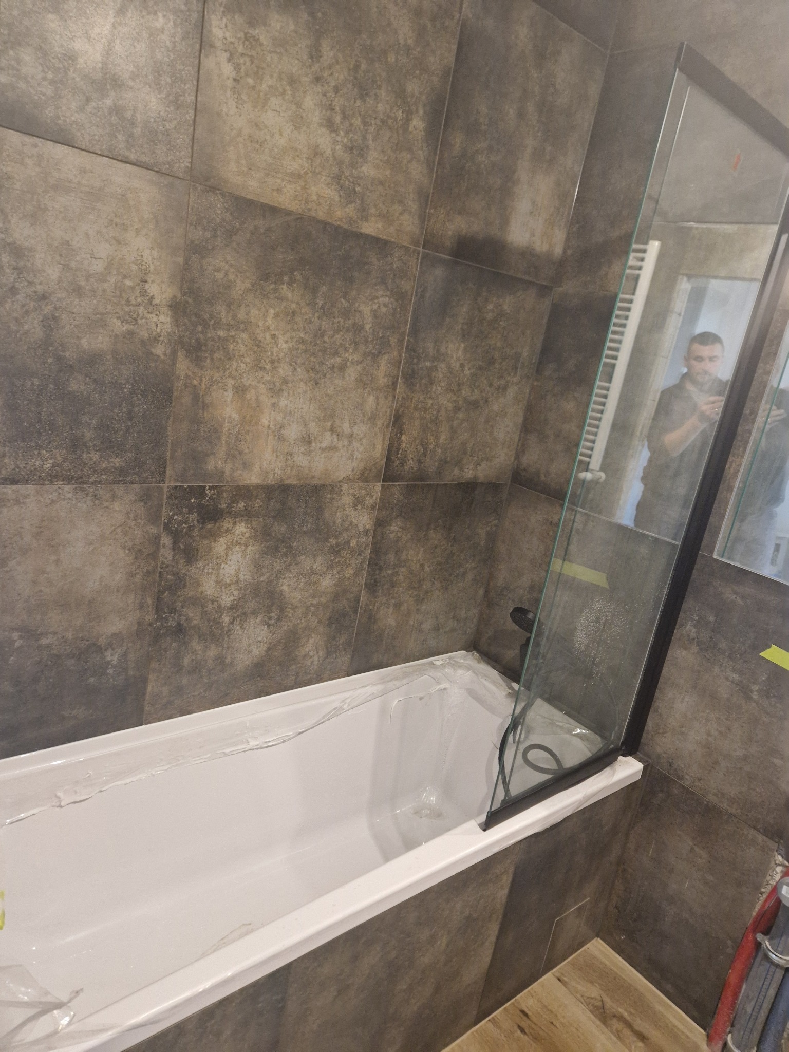 Nowoczesna łazienka z wanną i szklanym parawanem, wykończona płytkami w odcieniach szarości. Widoczne detale wykończeniowe i odbicie osoby w lustrze.