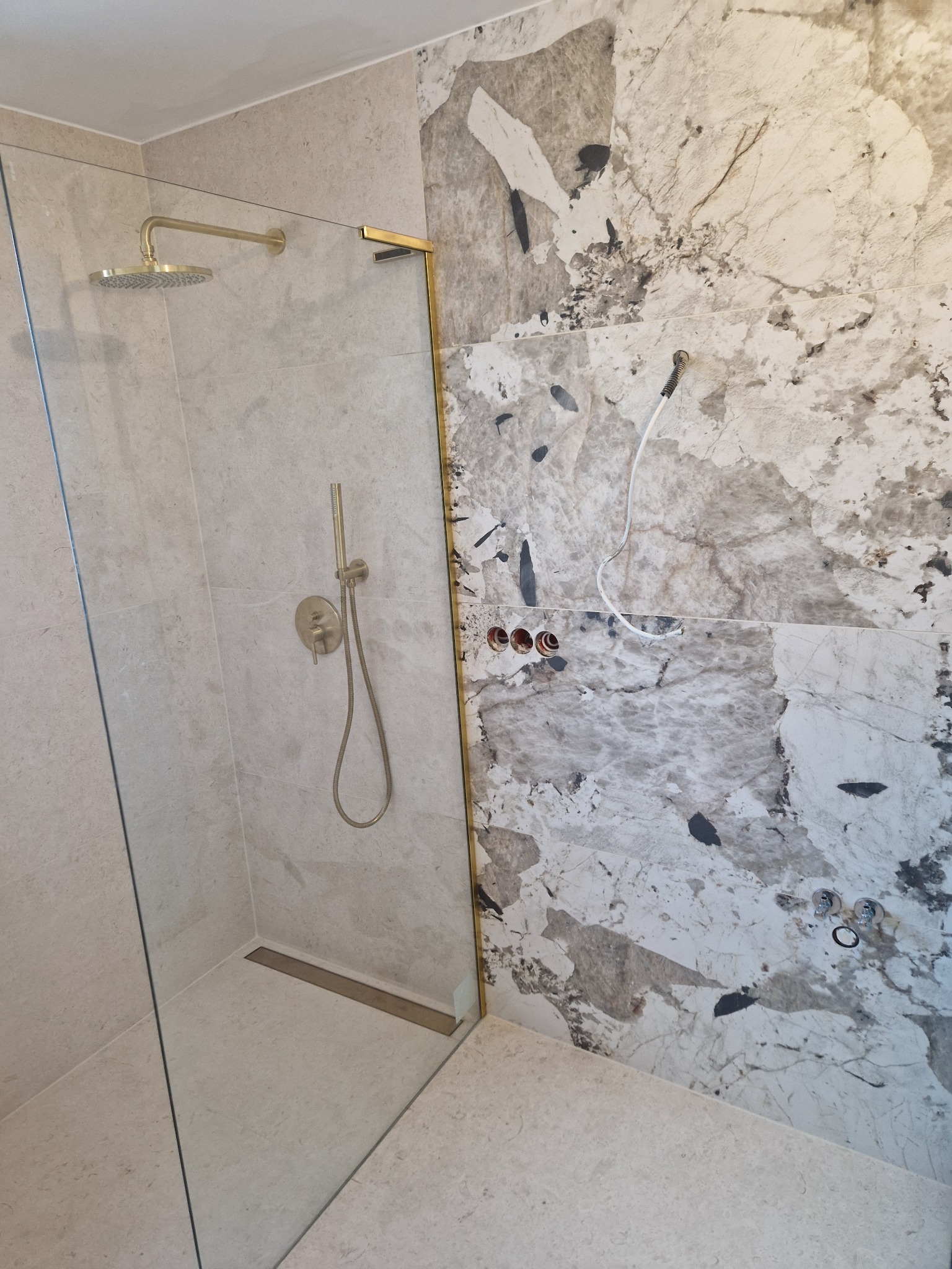 Kabina prysznicowa walk-in z złotymi detalami: deszczownica, bateria, rama. Ściany wyłożone płytami imitującymi marmur. Widoczne przygotowane przyłącza wodne.