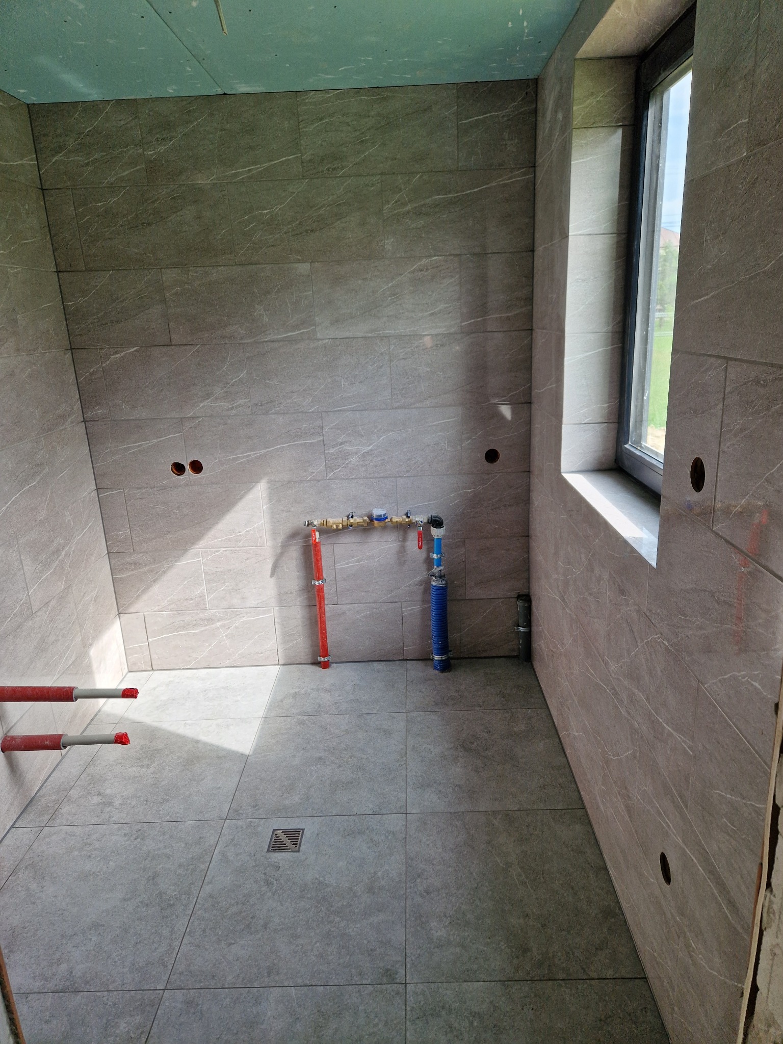 Łazienka w trakcie prac wykończeniowych: szare płytki imitujące marmur na ścianach i podłodze, widoczne instalacje wodne, okno z parapetem i otwory na punkty elektryczne.