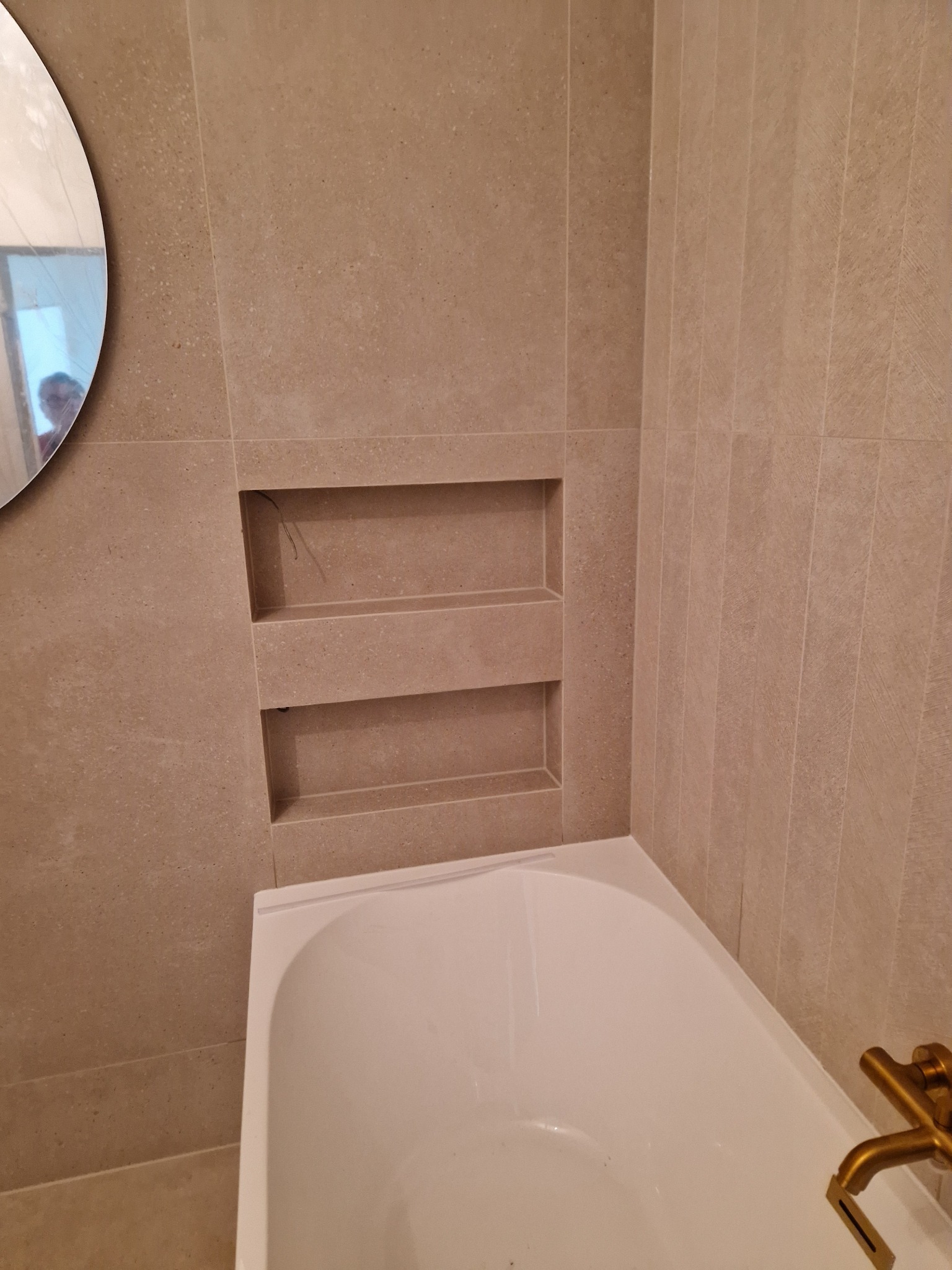 Beżowa łazienka z wanną i wnęką w ścianie wykończoną płytkami. Widoczne złote elementy armatury i okrągłe lustro.