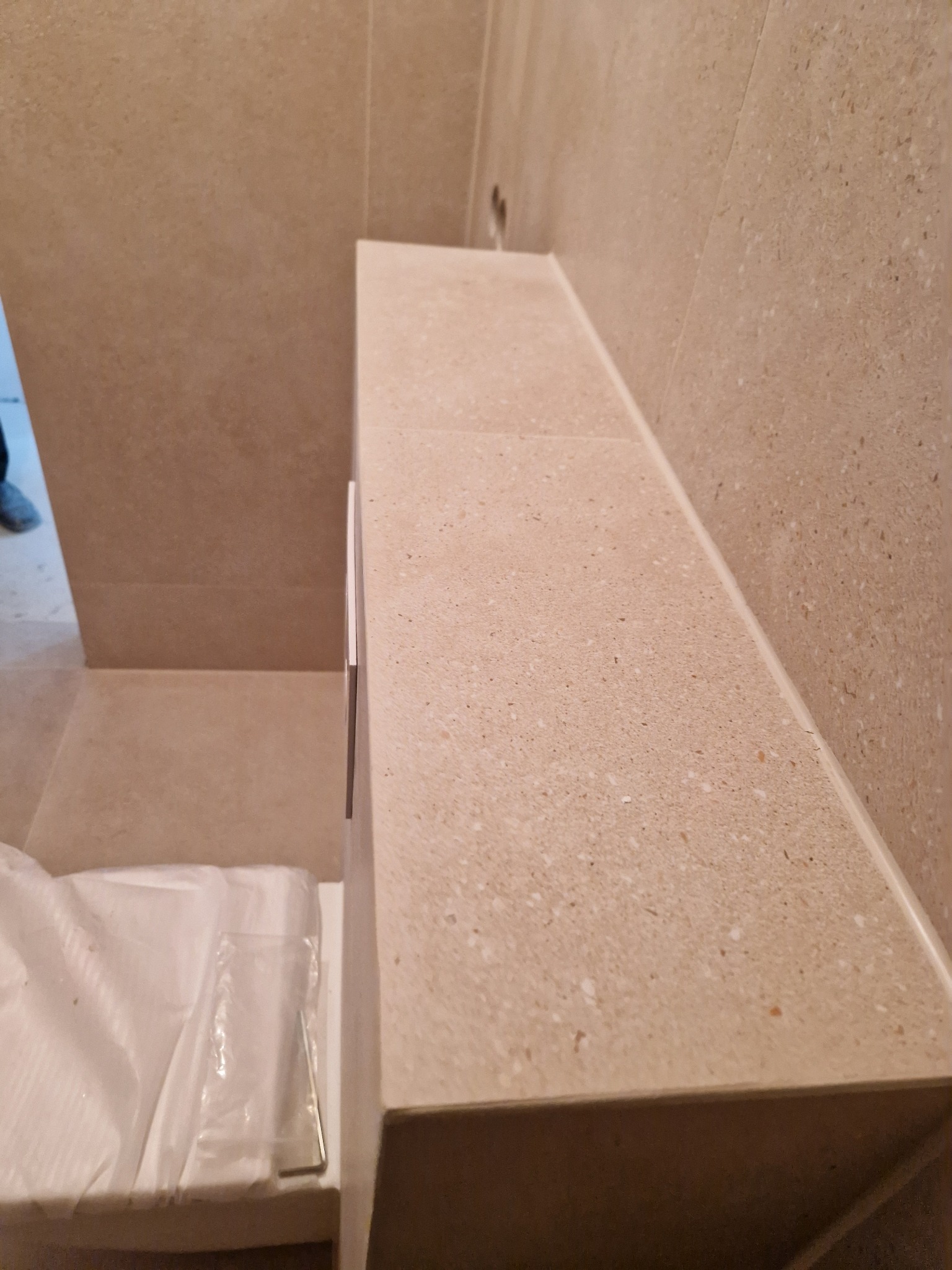 Beżowa, kamienna zabudowa w łazience z widocznymi fugami i fragmentem niezapakowanego wyposażenia. Minimalistyczny design, ciepłe oświetlenie.