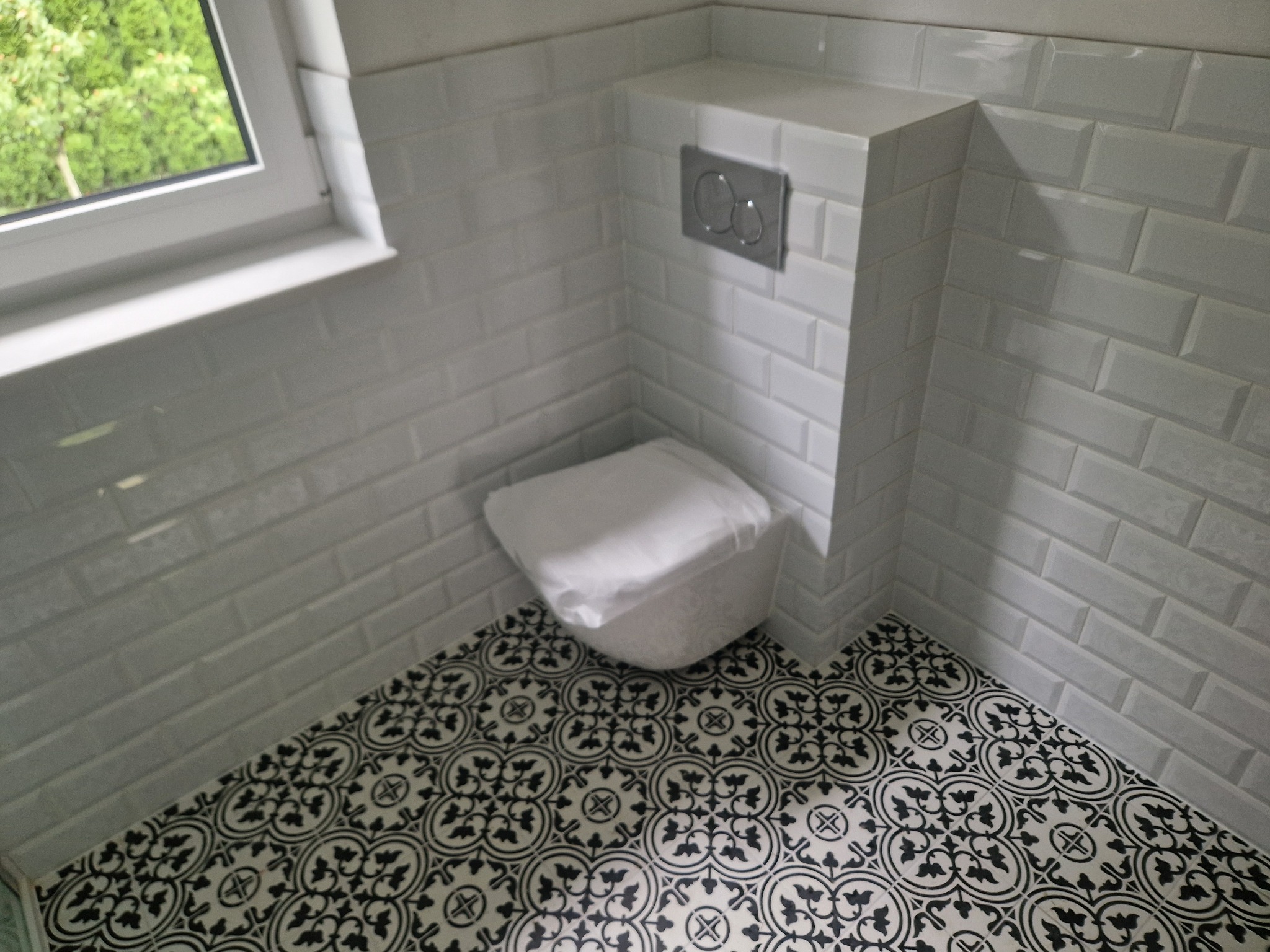 Biała łazienka z płytek typu metro i wzorzystą podłogą. Widoczny fragment okna. Toaleta zakryta folią ochronną, osprzęt chromowany.