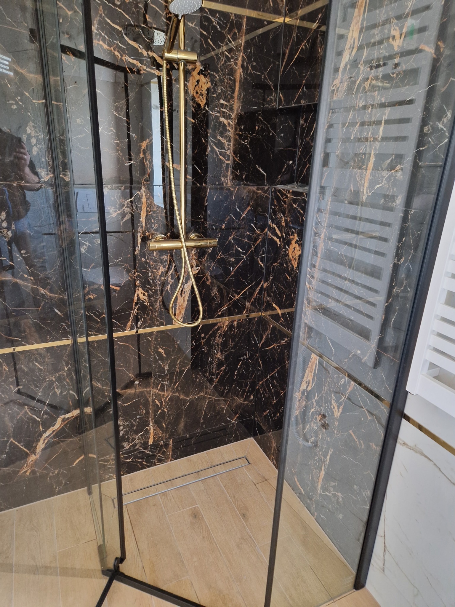 Kabina prysznicowa z czarnymi płytkami imitującymi marmur i złotą armaturą. Widoczny odpływ liniowy i grzejnik drabinkowy.