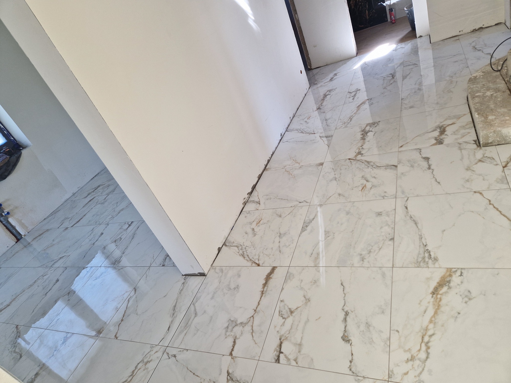 Błyszcząca, marmurowa podłoga w odcieniach bieli i beżu, świeżo ułożona w jasnym pomieszczeniu, z fragmentem białej ściany i widocznym progiem drzwi.