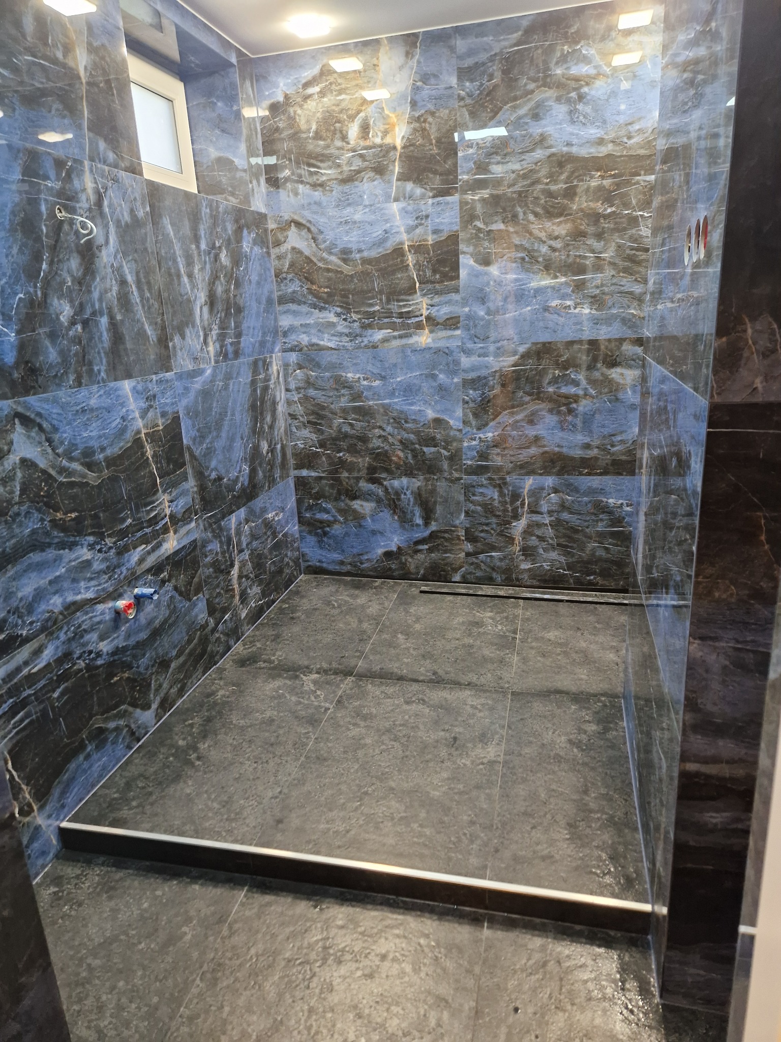 Kabina prysznicowa z ciemnymi, marmurkowymi płytkami na ścianach i podłodze. Widoczne przyłącza wodne i odpływ liniowy. W górze małe okno.