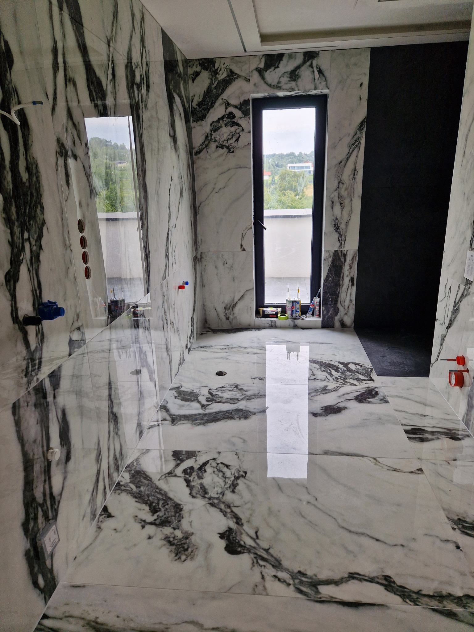 Łazienka w trakcie wykańczania: marmurowe płytki na ścianach i podłodze, okno z widokiem na zieleń, widoczne narzędzia i materiały budowlane na parapecie.