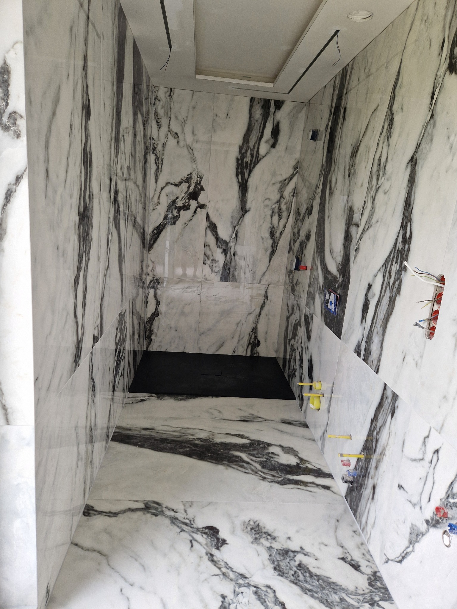 Wnętrze kabiny prysznicowej z marmurowymi płytkami w odcieniach bieli i czerni. Widoczne instalacje elektryczne i wodne przed ostatecznym wykończeniem.