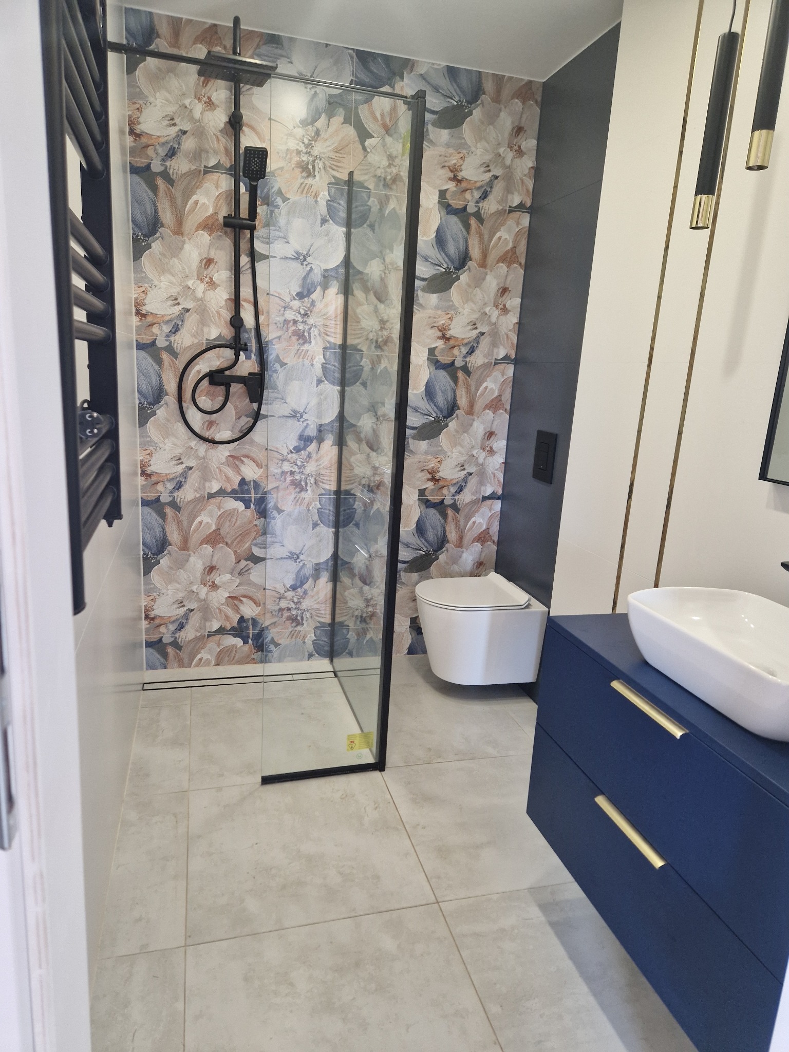 Nowoczesna łazienka z kabiną typu walk-in, płytkami w kwiaty, umywalką nablatową na szafce w kolorze granatowym i wiszącą toaletą. Oświetlenie punktowe.