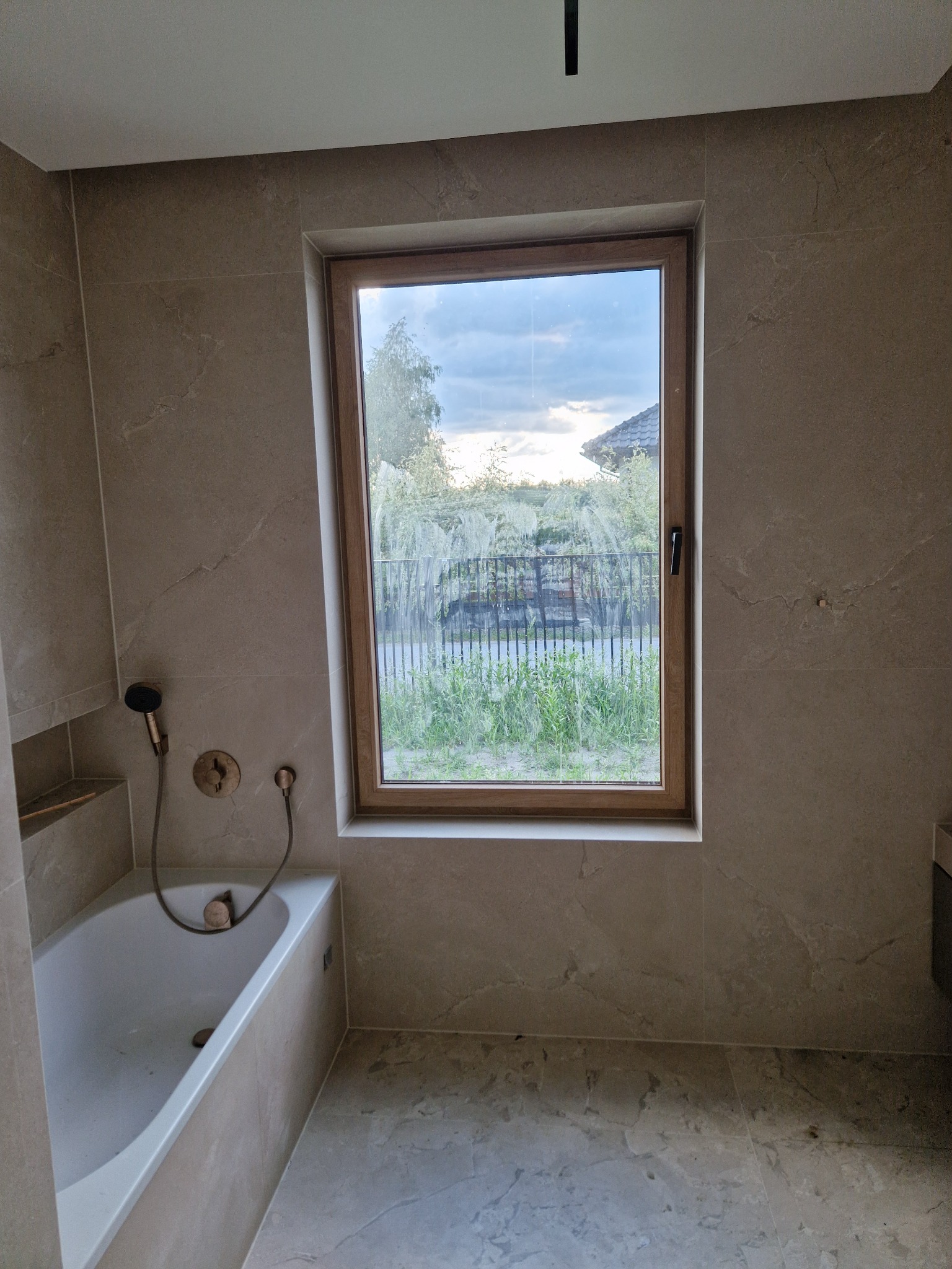 Wykończona łazienka z beżowymi płytkami imitującymi marmur, okno z widokiem na zieleń. Bateria prysznicowa w stylu retro. Minimalistyczny design.