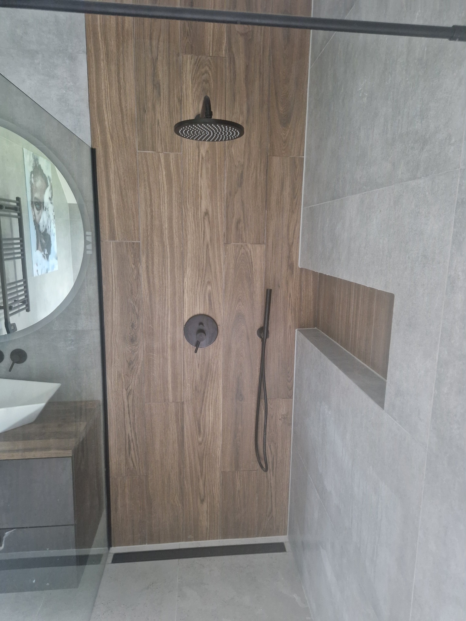 Nowoczesna kabina prysznicowa z deszczownicą i drewnopodobnymi płytkami, z wnęką w ścianie wykończoną szarą płytką. Widoczne elementy czarnej armatury.