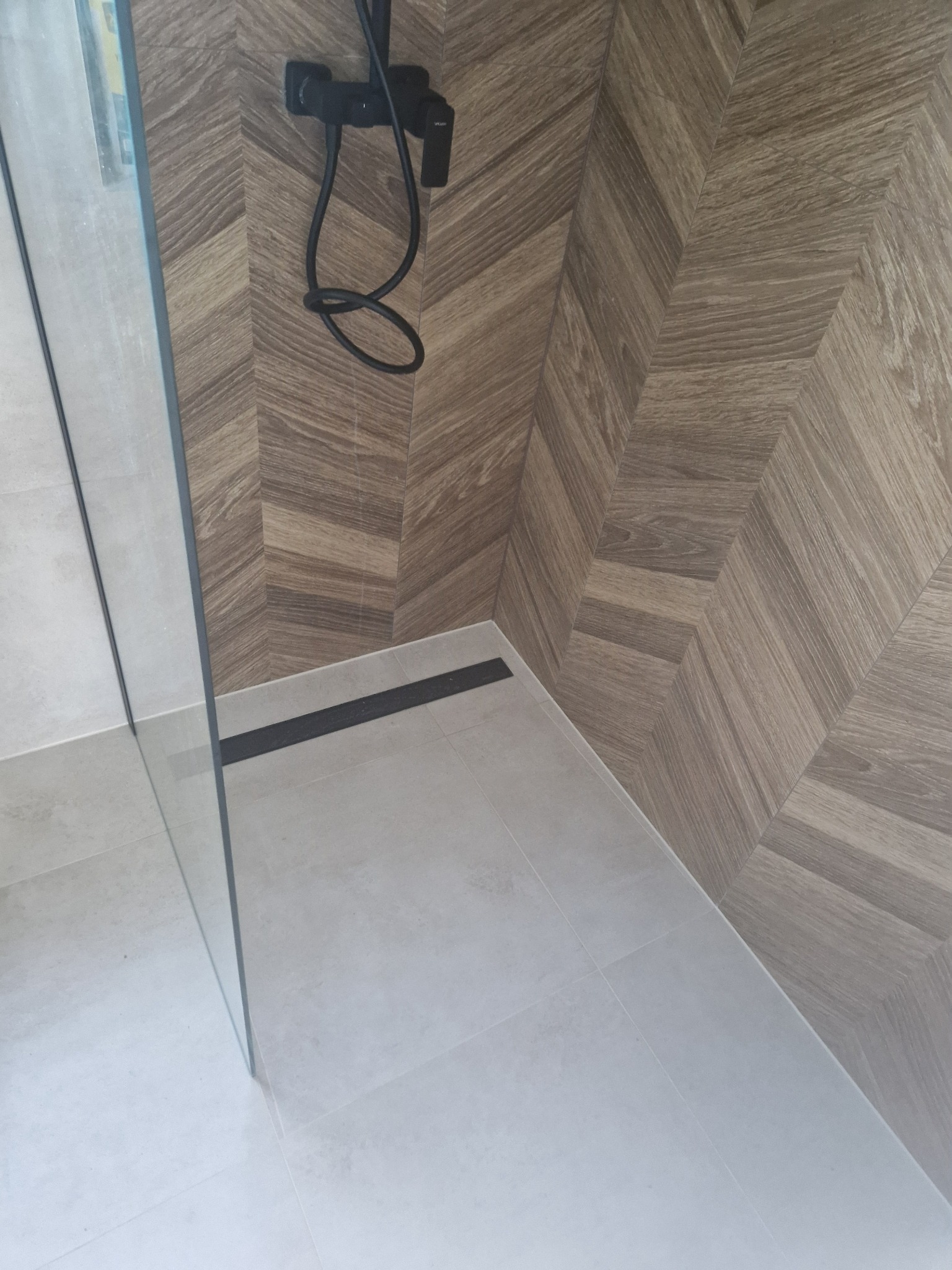 Nowoczesna kabina prysznicowa z drewnopodobną ścianą ułożoną w jodełkę i czarną armaturą. Widoczne szklane drzwi i odpływ liniowy.