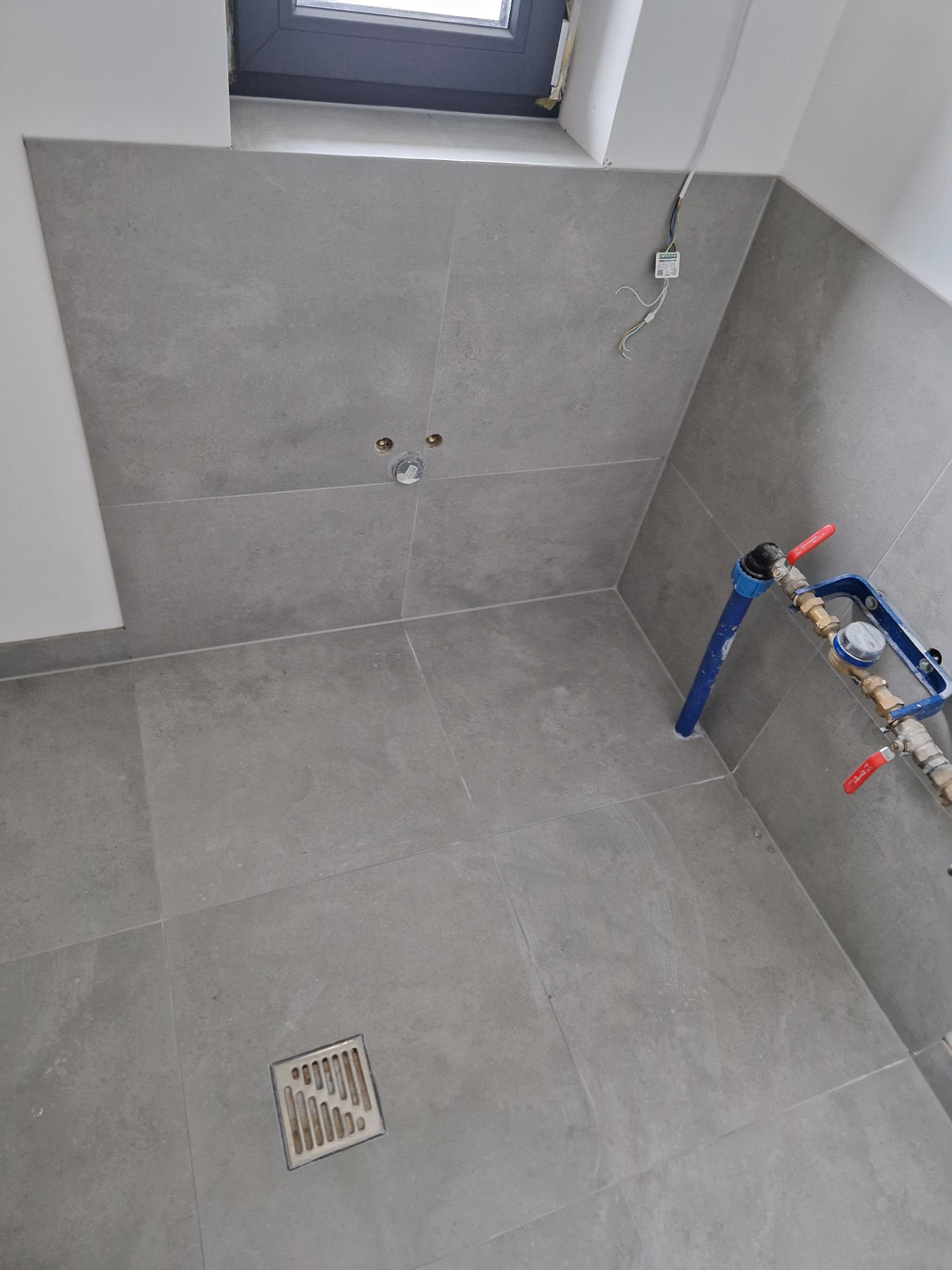 Szare płytki wielkoformatowe na ścianach i podłodze w łazience, widoczny odpływ liniowy, instalacja wodna i okno. Minimalistyczny design, surowy styl.