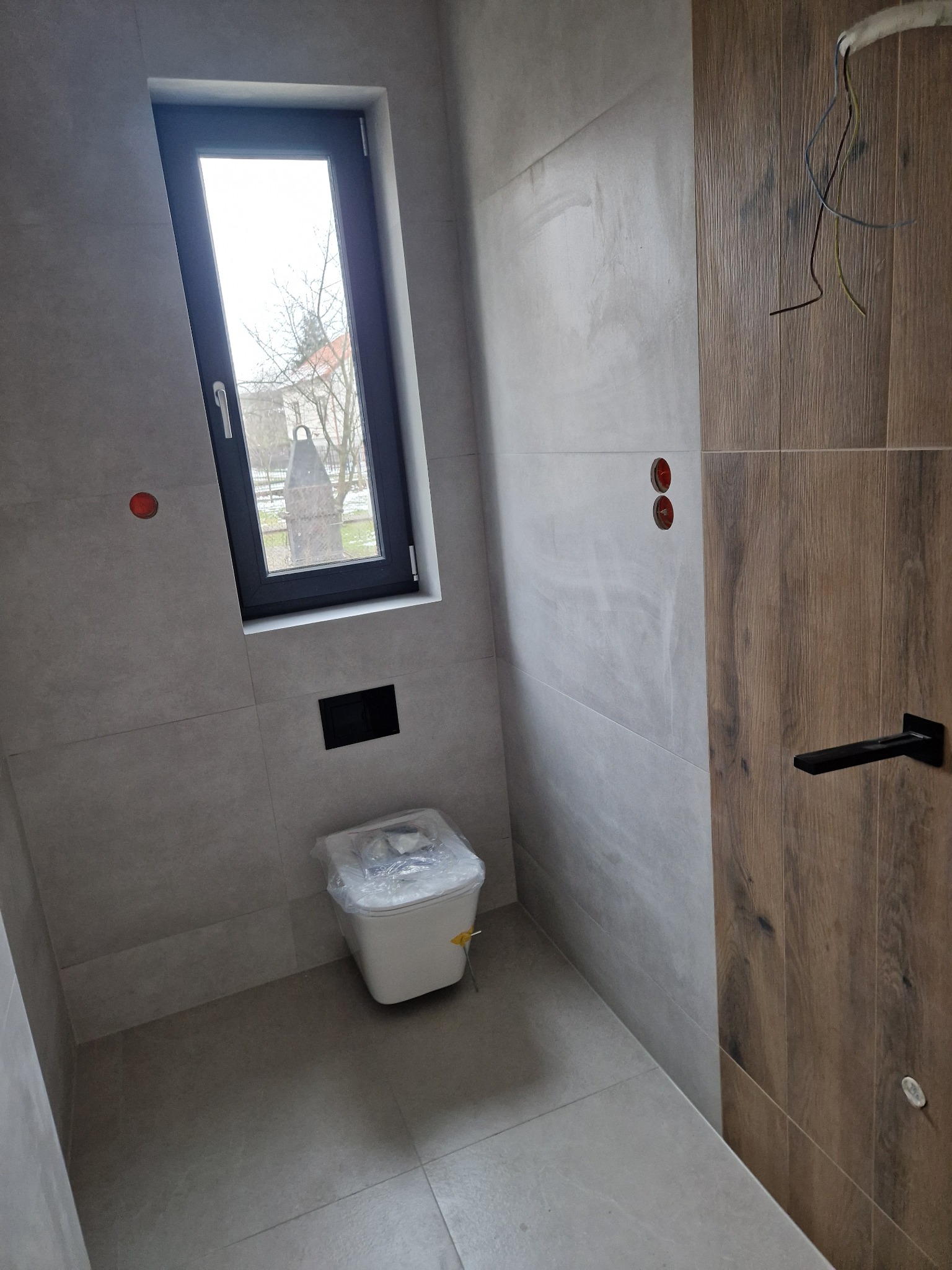 Nowoczesna łazienka w szarościach z akcentem drewna. Ukończona instalacja podtynkowa WC, okno z widokiem na zewnątrz. Częściowo widoczne przewody elektryczne.