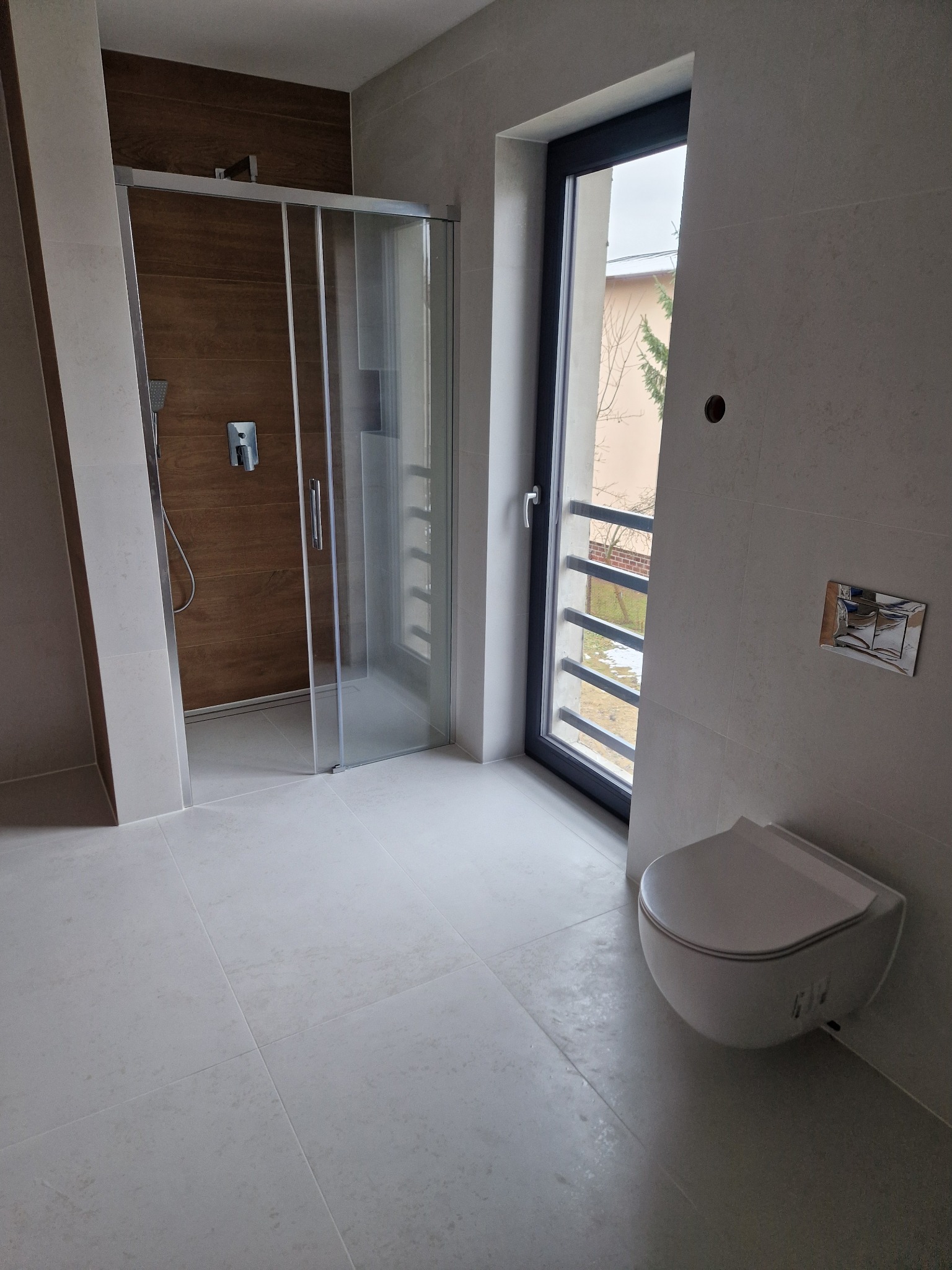 Nowoczesna łazienka z kabiną prysznicową typu walk-in, podwieszaną toaletą i oknem z widokiem na zewnątrz. Płytki w odcieniach szarości i brązu.