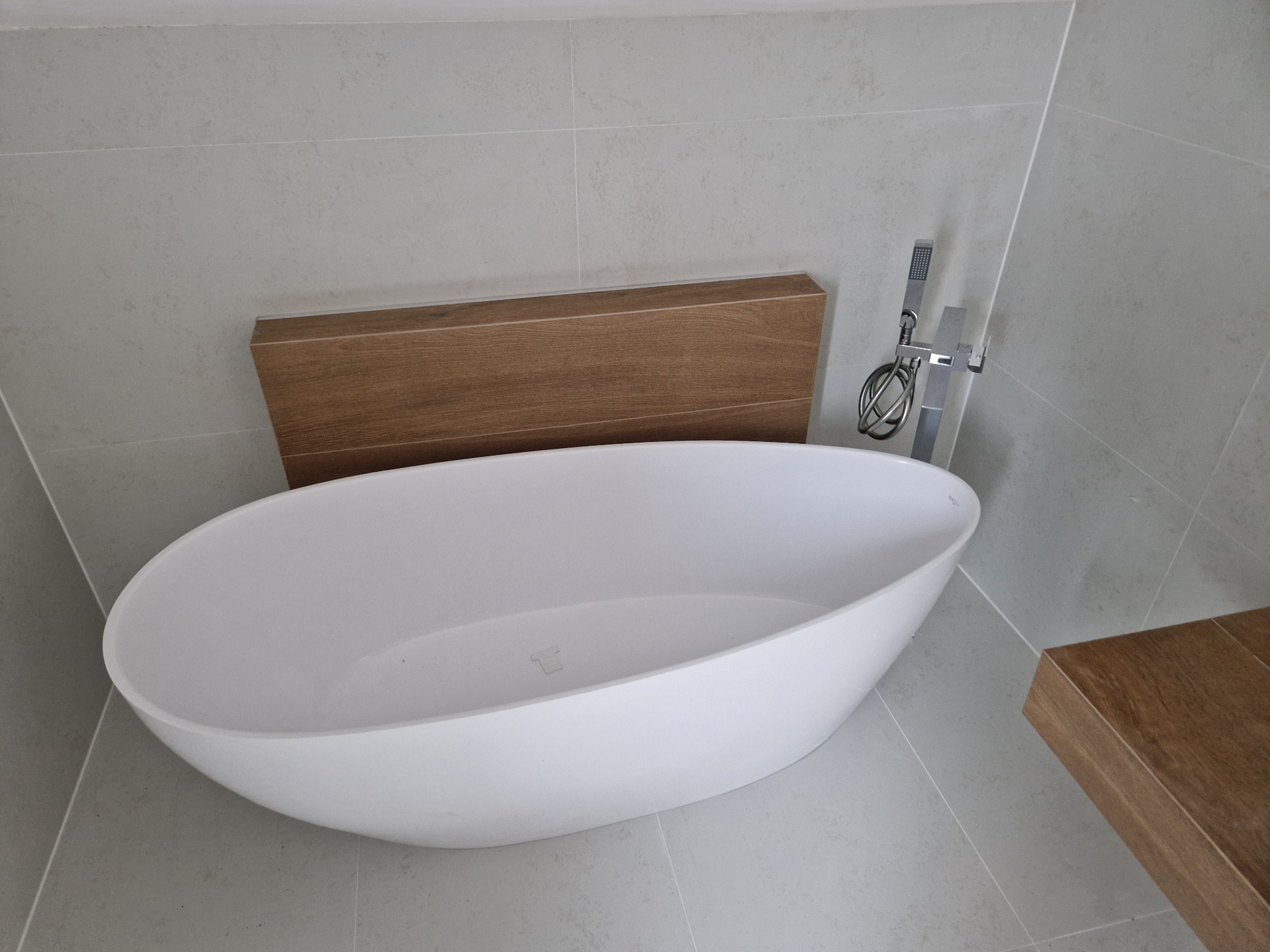 Nowoczesna, owalna wanna wolnostojąca w jasnej łazience z szarymi płytkami i drewnianymi akcentami. Minimalistyczna armatura.