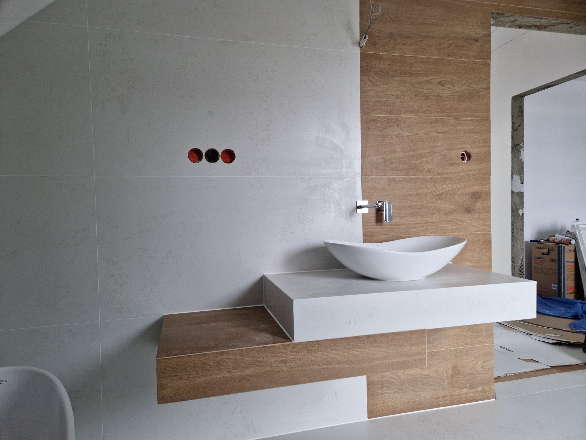 Nowoczesna łazienka w trakcie wykańczania: biała umywalka na blacie, płytki imitujące drewno i szare płytki ścienne z widocznymi otworami na gniazdka.