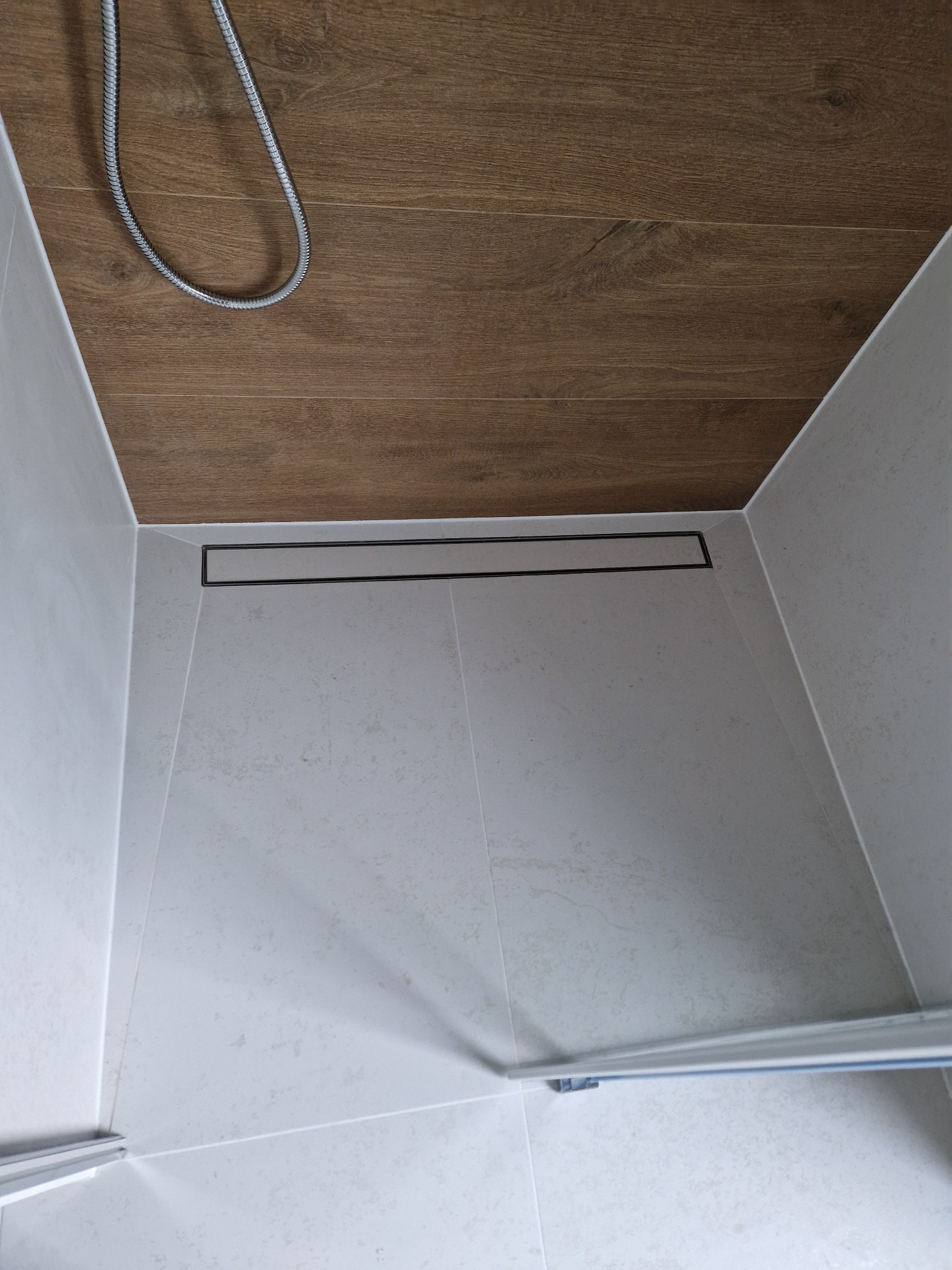 Nowoczesny prysznic walk-in z odpływem liniowym, wykończony płytkami w odcieniach bieli i brązu. Widoczna słuchawka prysznicowa i fragment szklanej ścianki.