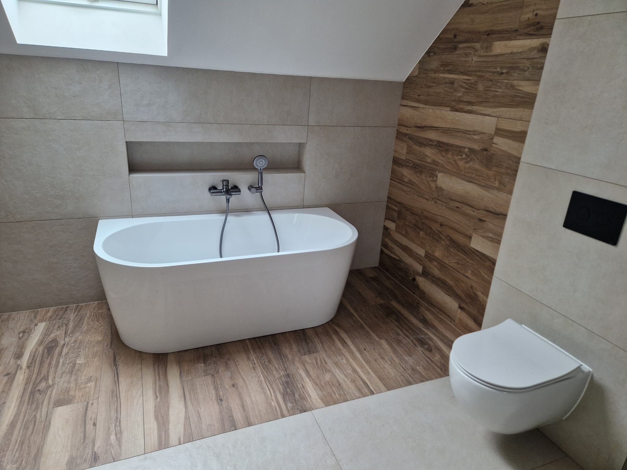Nowoczesna łazienka z wolnostojącą wanną, podwieszaną toaletą i płytkami imitującymi drewno na podłodze i ścianie. W ścianie wnęka z baterią wannową.